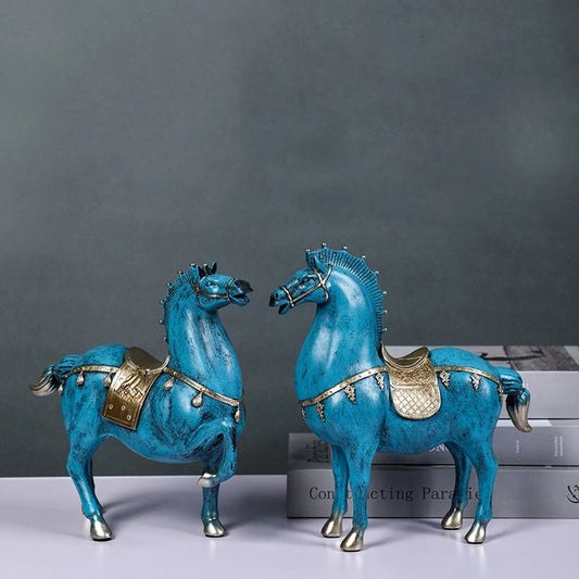 Pair of blue horse statues with gold saddles and details, standing on a table with books in the background.