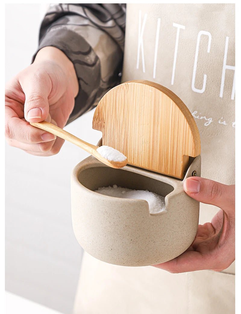 Person holding a ceramic seasoning jar with a wooden lid, while another hand holds a wooden spoon filled with sugar