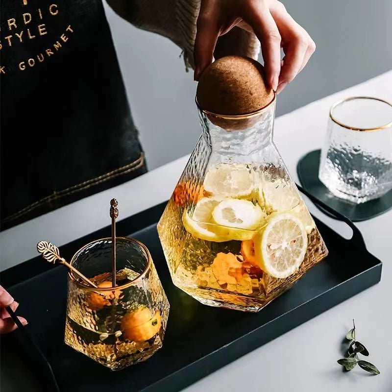 A person touching the lid of a Nordic glass pitcher filled with water and lemon slices, with two cups beside it, one filled with lemon water and the other empty.